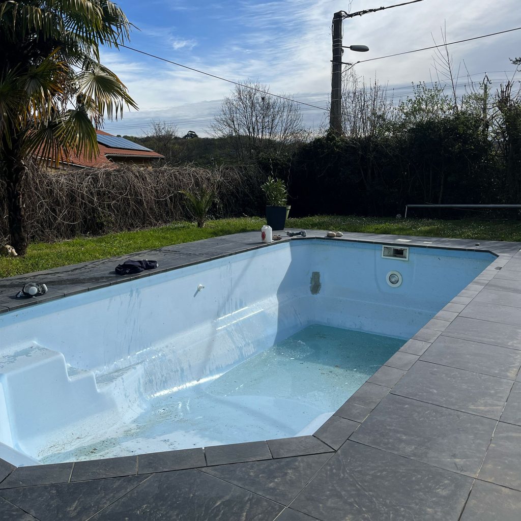 Piscine extérieure rectangulaire partiellement remplie d'eau, entourée de dalles en pierre, avec un jardin et des arbres en arrière-plan sous un ciel clair.